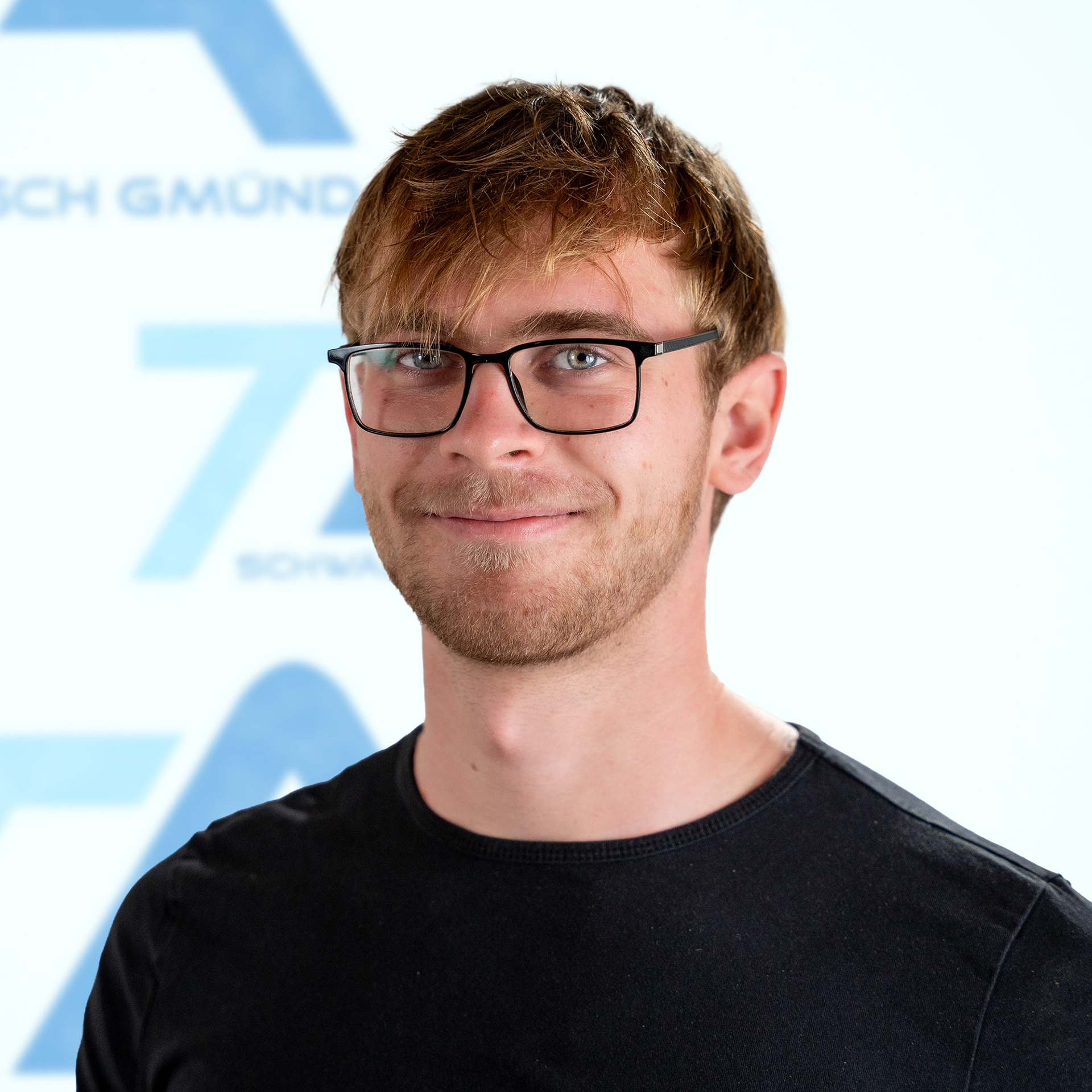 Ein junger Mann mit kurzen braunen Haaren und Brille, der ein schwarzes T-Shirt trägt.