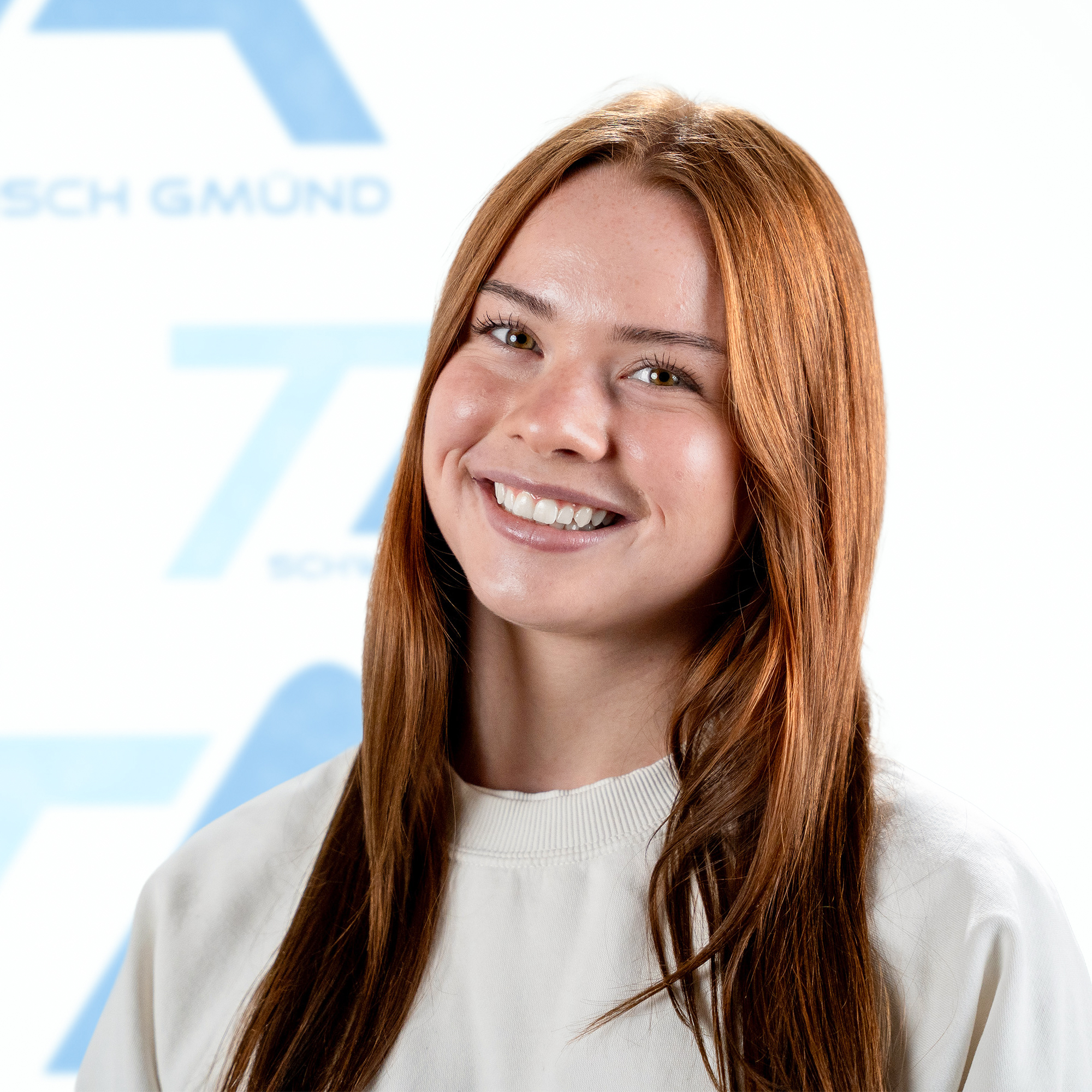 Eine junge Frau mit langen rotbraunen Haaren, die vor einem Hintergrund mit Markenlogo in die Kamera lächelt.
