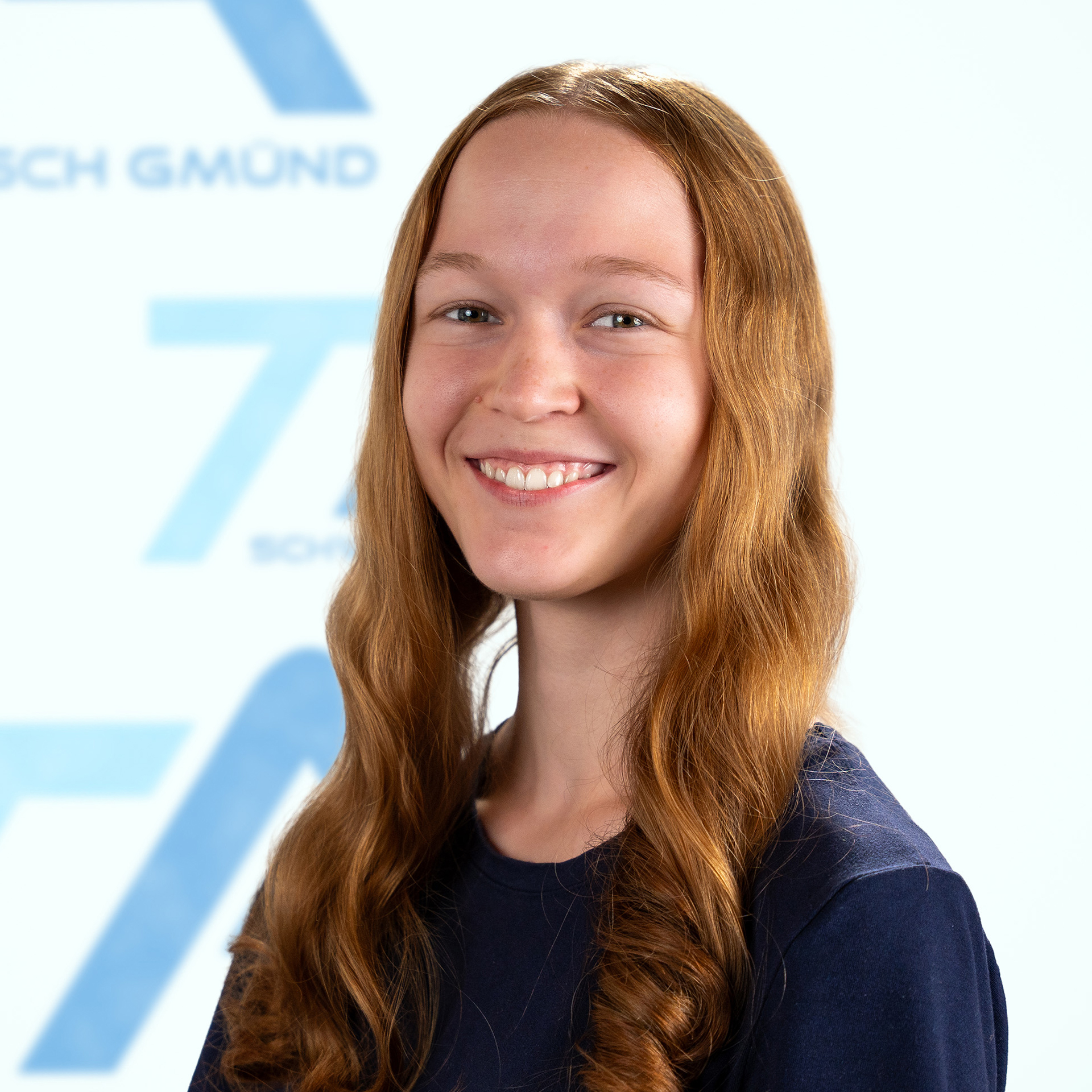 Eine lächelnde junge Frau mit langen rotbraunen Haaren steht vor einem verschwommenen weiß-blauen Logo.